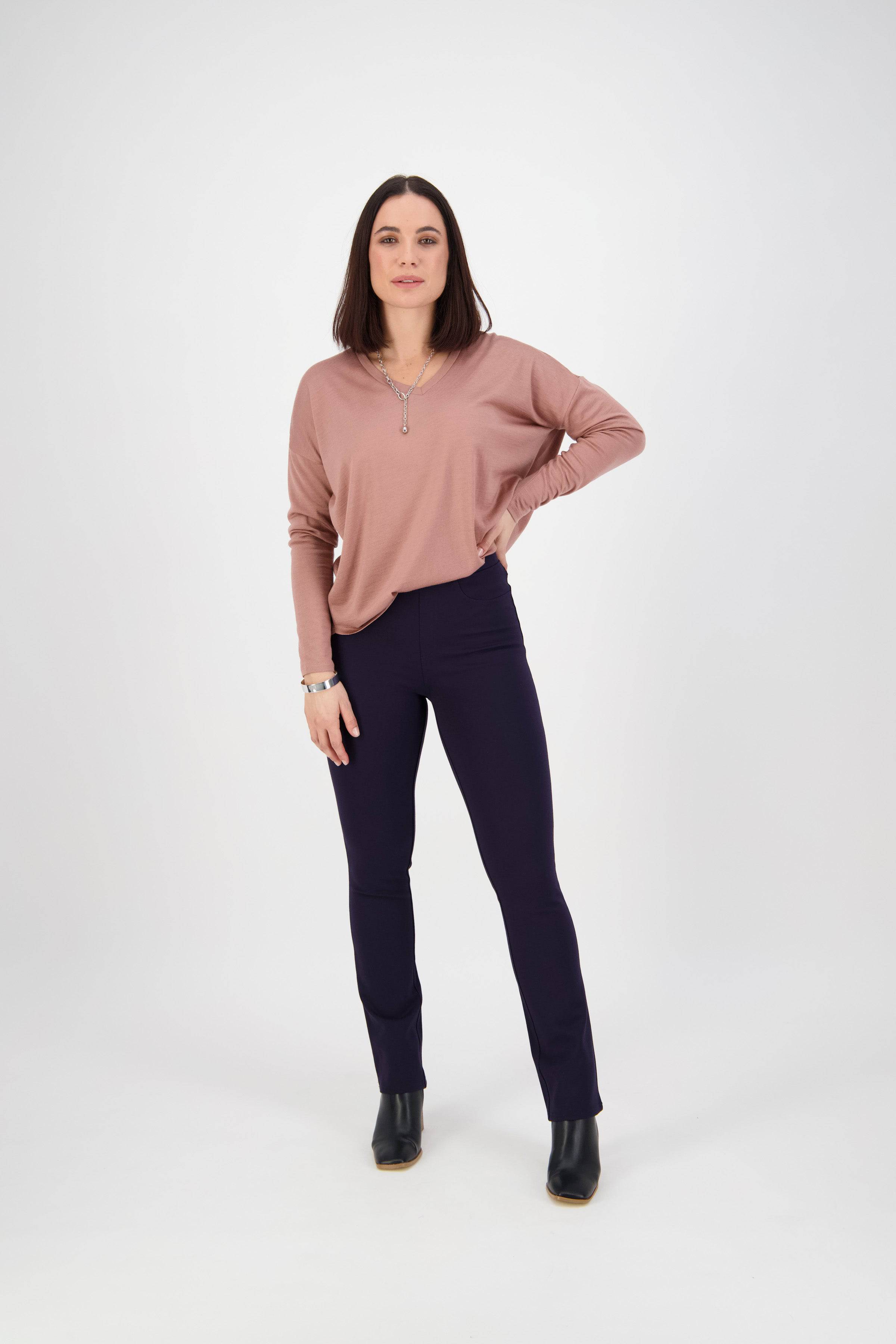 A woman with straight brown hair stands against a white background wearing a pink top, black ankle boots, and Vassalli Bay Ponti Pull On - Navy pants. She poses with one hand on her hip, looking forward.