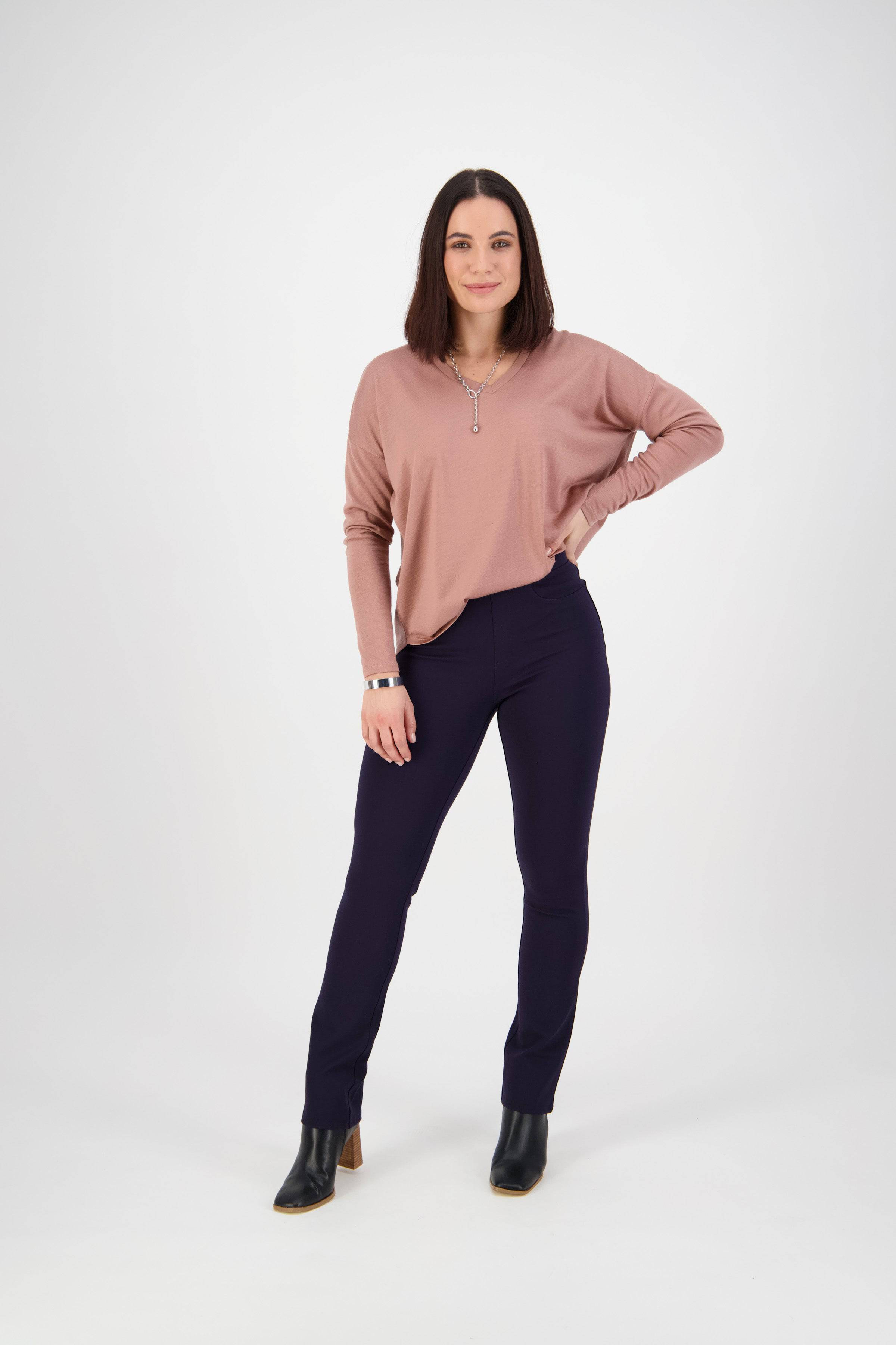 A woman with straight, shoulder-length brown hair stands confidently against a white background, wearing a mauve long-sleeve top, Vassalli Bay Ponti Pull On pants in navy, and black ankle boots.