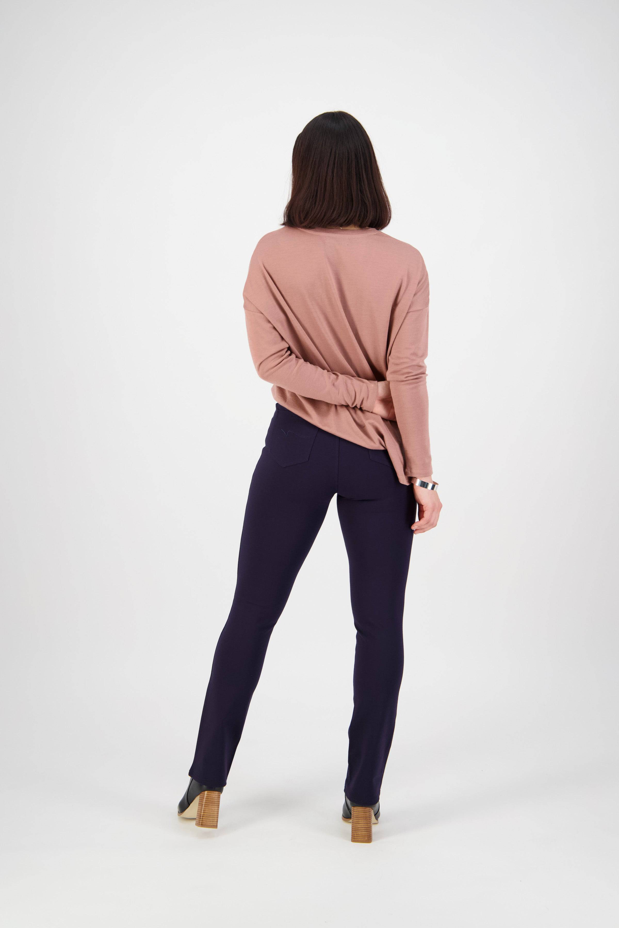 A woman with straight dark hair stands facing away, wearing Vassalli's Bay Ponti Pull On slim leg pants in navy and a mauve long-sleeve top. One arm is bent behind her back against a plain white background.