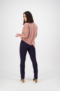 A woman with shoulder-length dark hair faces away, looking over her shoulder. She wears a loose pink top, Vassalli Bay Ponti Pull On pants in navy, and heeled boots against a plain white background.