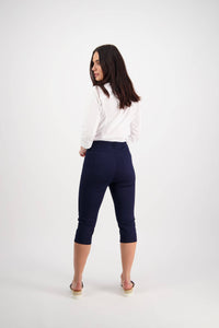 A woman with long dark hair faces away, wearing a white long-sleeve shirt, Vassalli's Atlas 3/4 Pant in navy with an elastic waistband, and beige sandals against a plain white background.