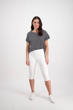 A woman with long brown hair stands confidently against a white background, wearing Vassalli's Atlas 3/4 Pant in white, a black and white striped top, and white sneakers.
