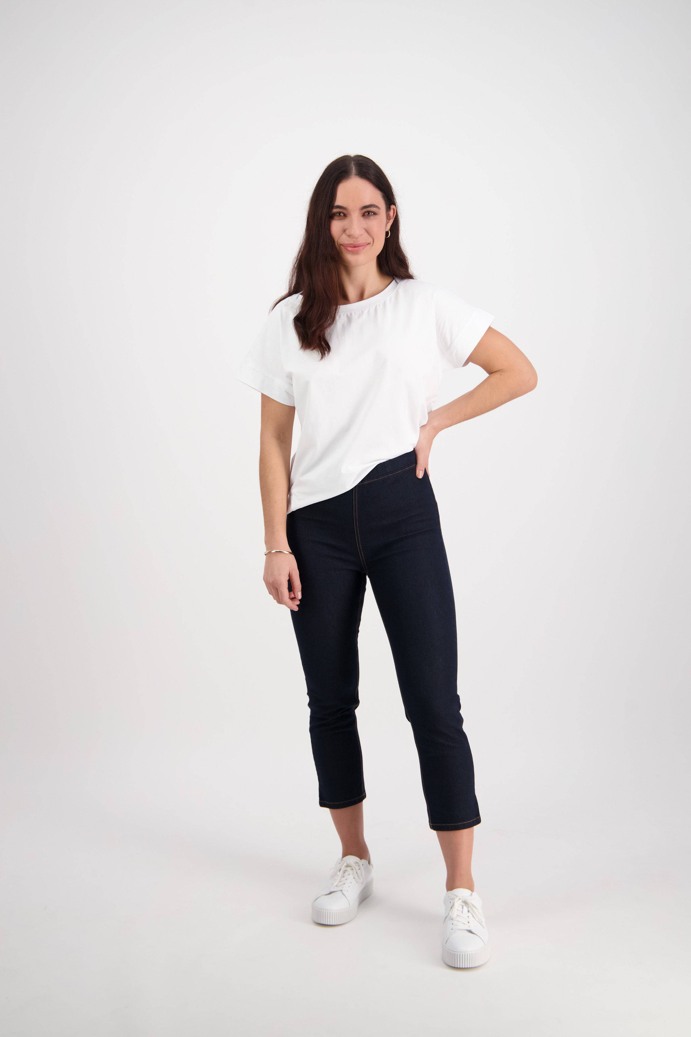 A woman with long brown hair stands against a white background, wearing a white t-shirt and Vassalli's Harbour 7/8 Pant in Indigo Denim (Contrast Stitch), paired with white sneakers. She smiles and rests one hand on her hip.