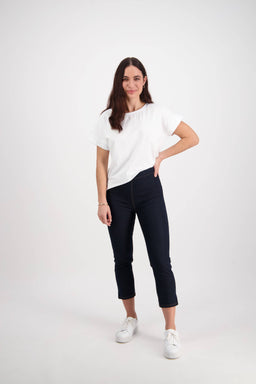 A woman with long brown hair stands against a white background, wearing a white t-shirt and Vassalli's Harbour 7/8 Pant in Indigo Denim (Contrast Stitch), paired with white sneakers. She smiles and rests one hand on her hip.