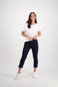 A young woman with long brown hair, wearing a white t-shirt and Vassalli Harbour 7/8 Pant - Indigo Denim (Contrast Stitch), smiles in a bright studio with a plain white background.
