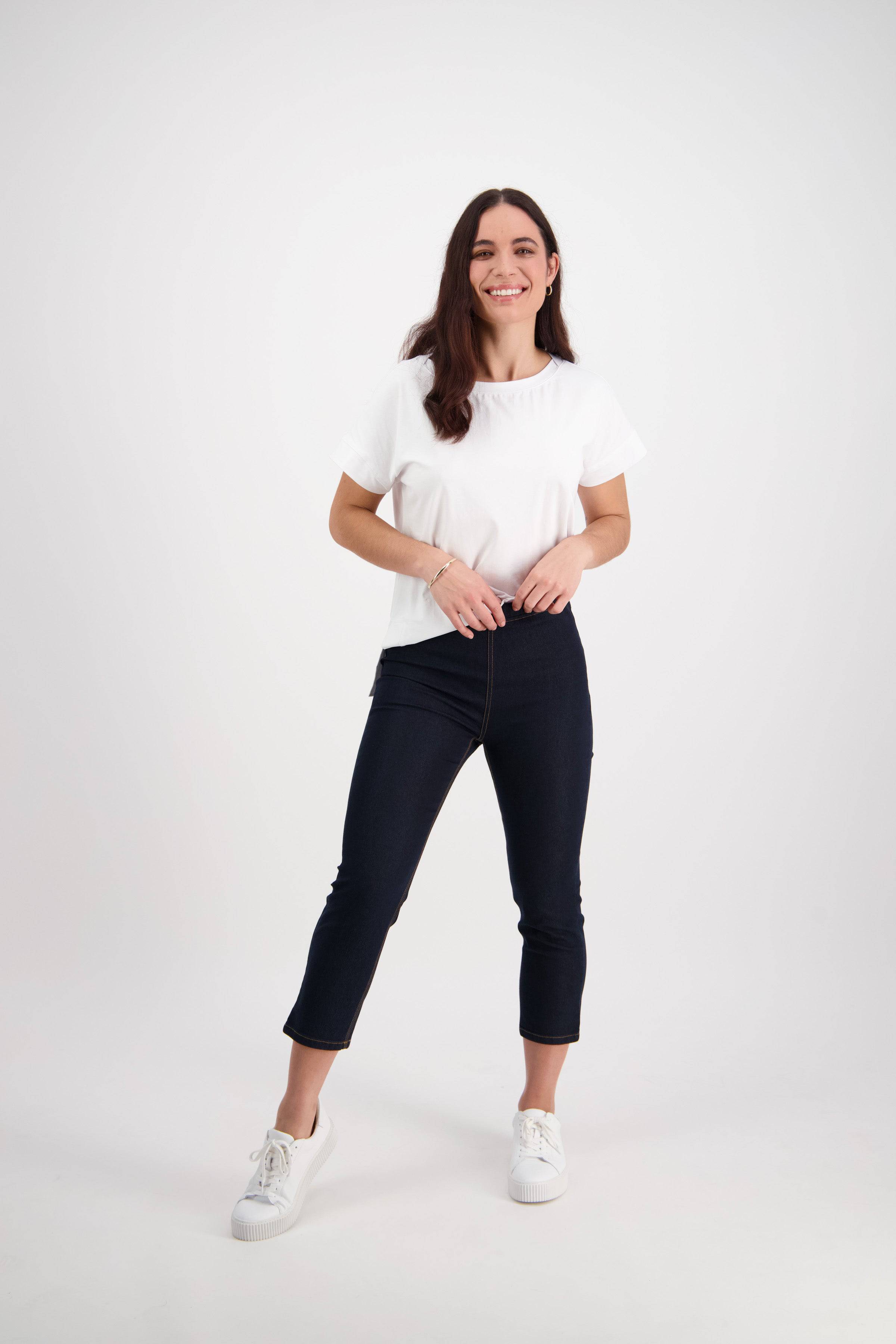 A young woman with long brown hair, wearing a white t-shirt and Vassalli Harbour 7/8 Pant - Indigo Denim (Contrast Stitch), smiles in a bright studio with a plain white background.