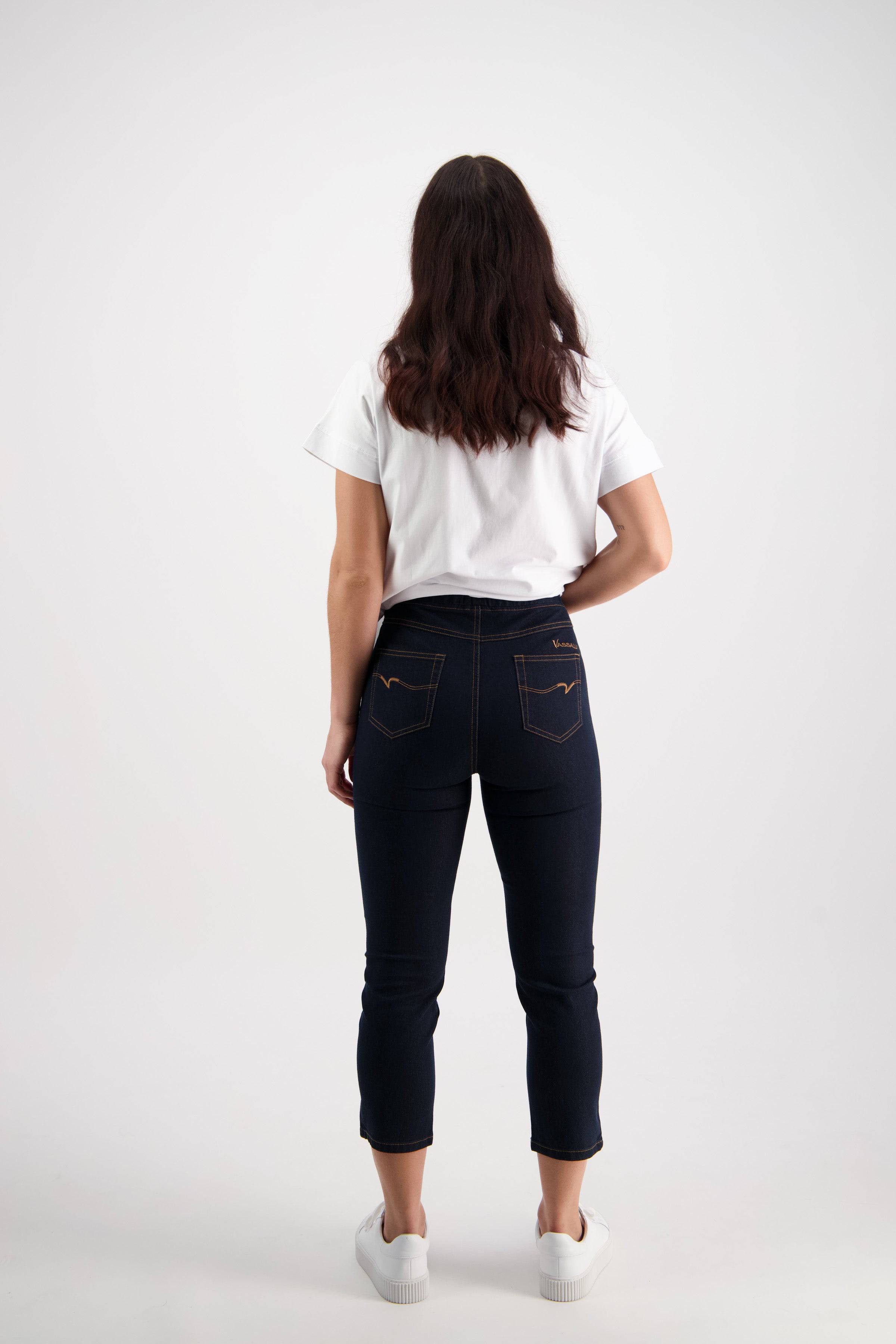 A person with long brown hair, in a white t-shirt and Vassalli Harbour 7/8 Pant - Indigo Denim (Contrast Stitch), wearing white sneakers, stands facing away from the camera against a plain white background.