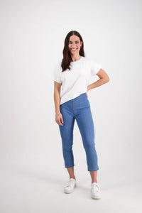 A woman with long brown hair smiles against a white background, wearing a white t-shirt, white sneakers, and the Vassalli Harbour 7/8 Pant in Light Denim (Contrast Stitch), one hand on her hip and the other at her side.