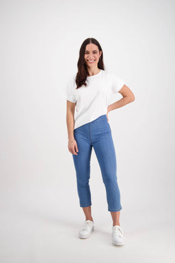 A woman with long brown hair smiles against a white background, wearing a white t-shirt, white sneakers, and the Vassalli Harbour 7/8 Pant in Light Denim (Contrast Stitch), one hand on her hip and the other at her side.