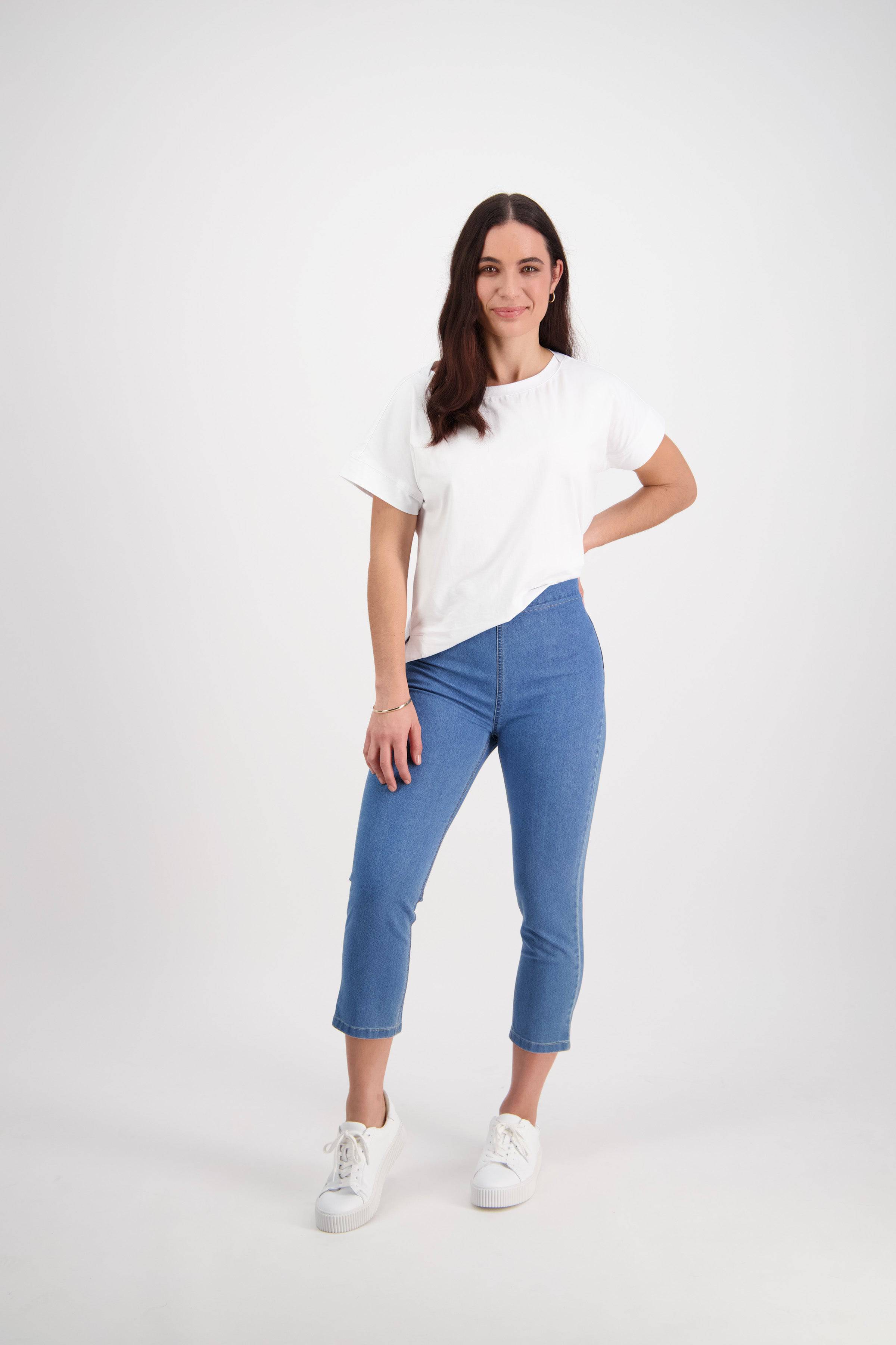 A woman with long brown hair smiles against a white background, wearing a white t-shirt, Vassalli Harbour 7/8 Pant - Light Denim (Contrast Stitch), and white sneakers.