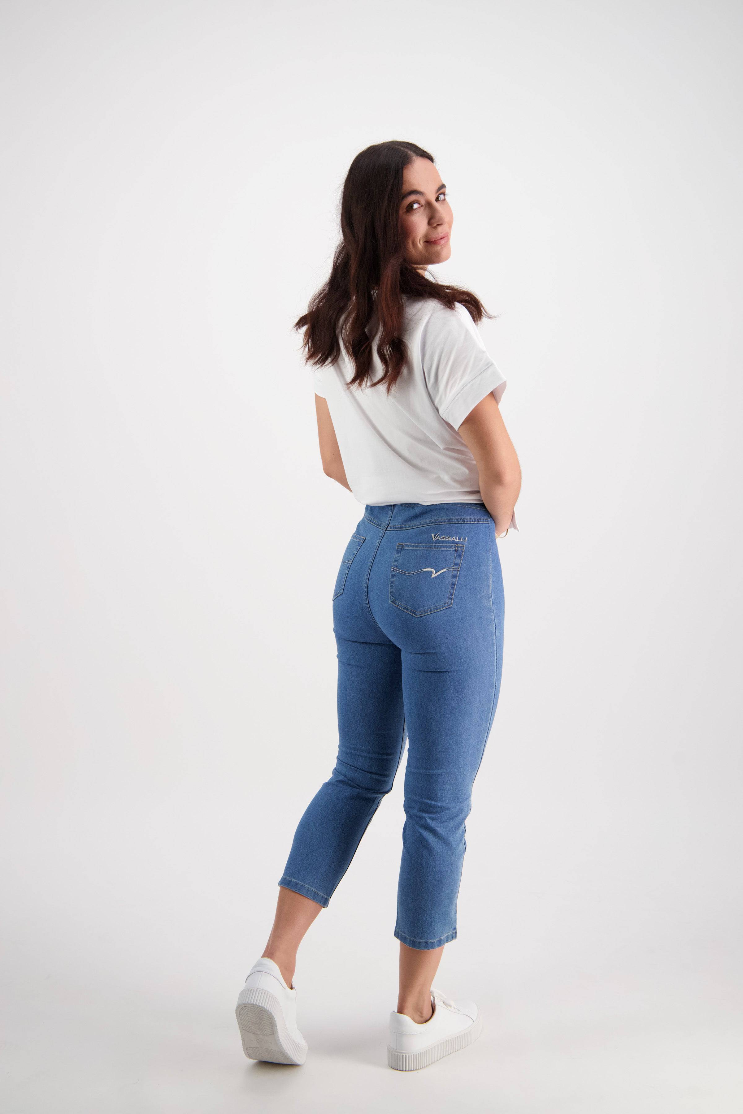 A woman with long dark hair, smiling and looking over her shoulder, wears the Vassalli Harbour 7/8 Pant - Light Denim (Contrast Stitch) and a white t-shirt in a simple white studio setting.