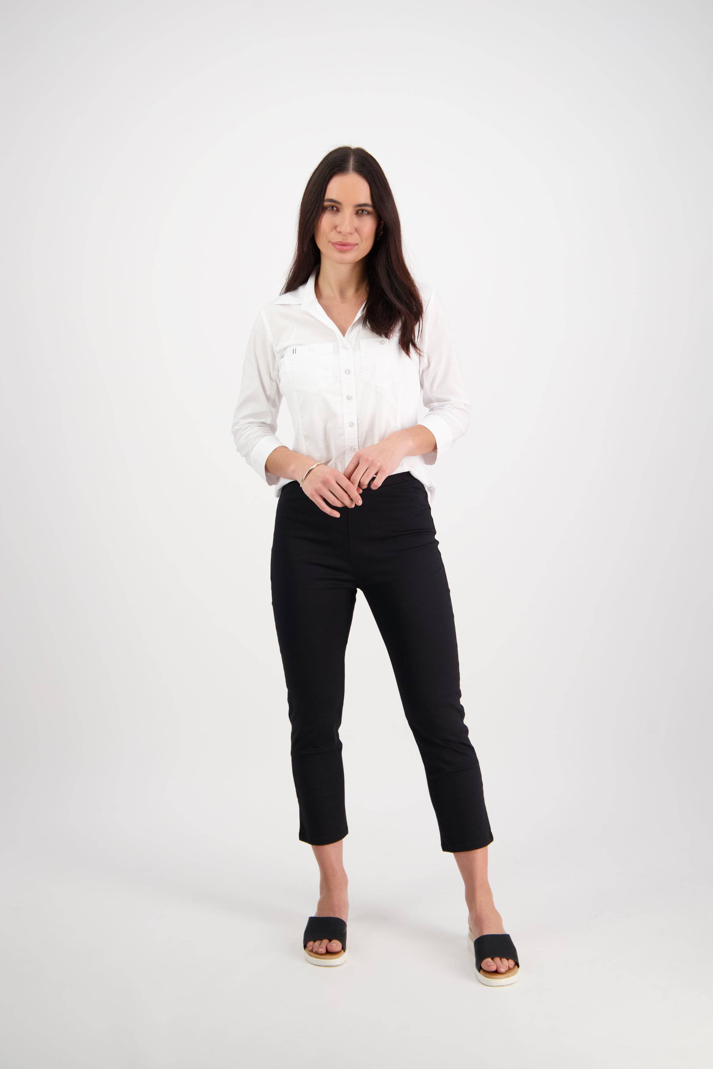 A woman with long dark hair stands against a plain white background wearing the Vassalli Harbour 7/8 Pant in black, a stretch skinny-leg style, paired with a white button-up shirt and black open-toe sandals; her hands rest near her waist.