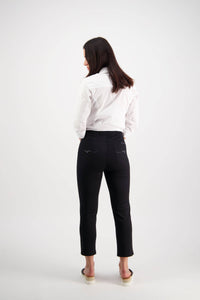A woman with straight dark hair, barefoot and facing away from the camera, wears a white long-sleeve shirt and Vassalli Harbour 7/8 Pant - Black against a plain white background.