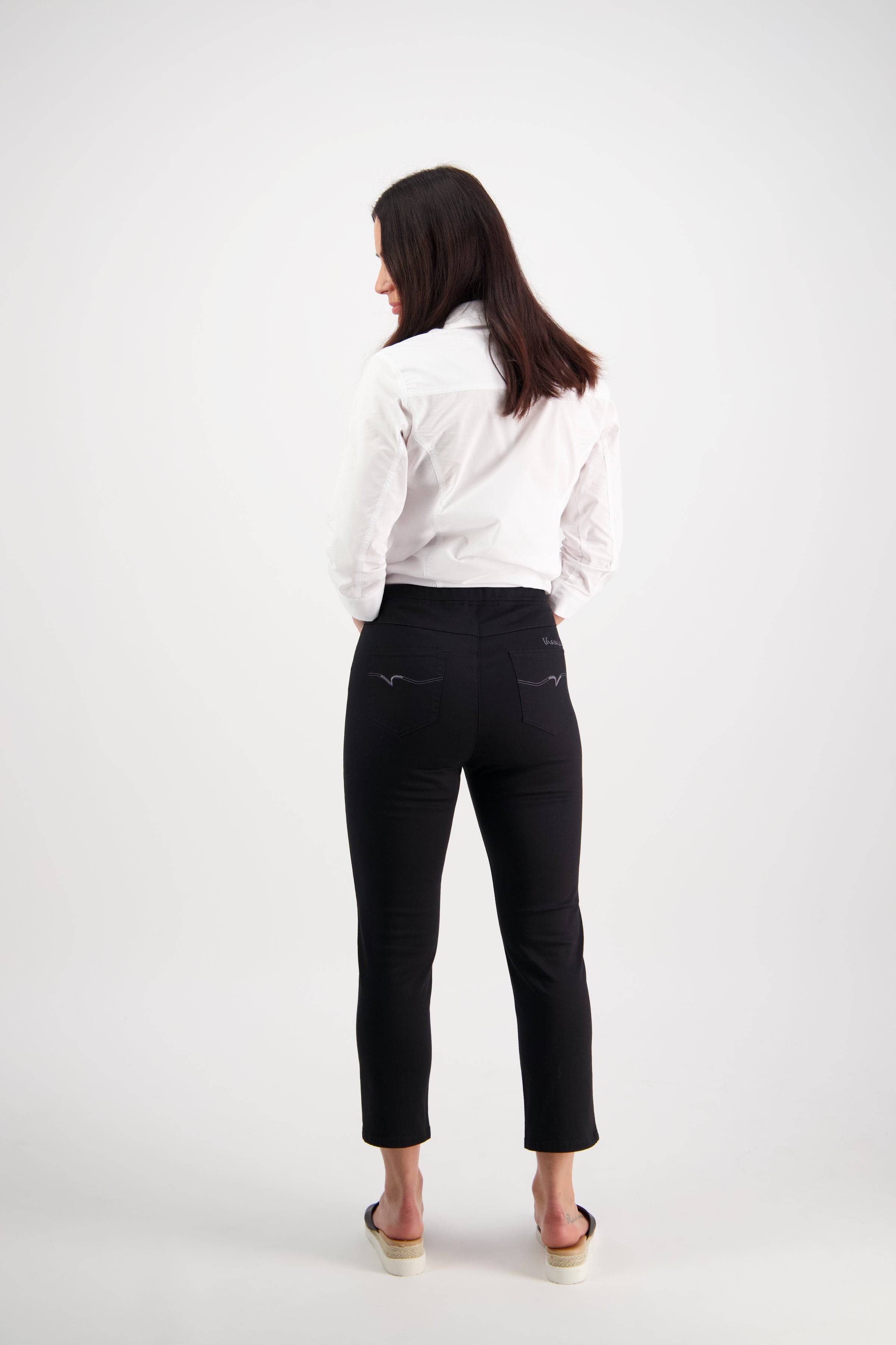 A woman with straight dark hair, barefoot and facing away from the camera, wears a white long-sleeve shirt and Vassalli Harbour 7/8 Pant - Black against a plain white background.