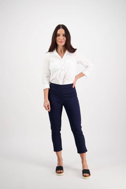 A woman with long dark hair stands against a white background, smiling with one hand on her hip. She wears a white cotton stretch button-up shirt and Vassalli Harbour 7/8 Pant in Navy, paired with black open-toed shoes.