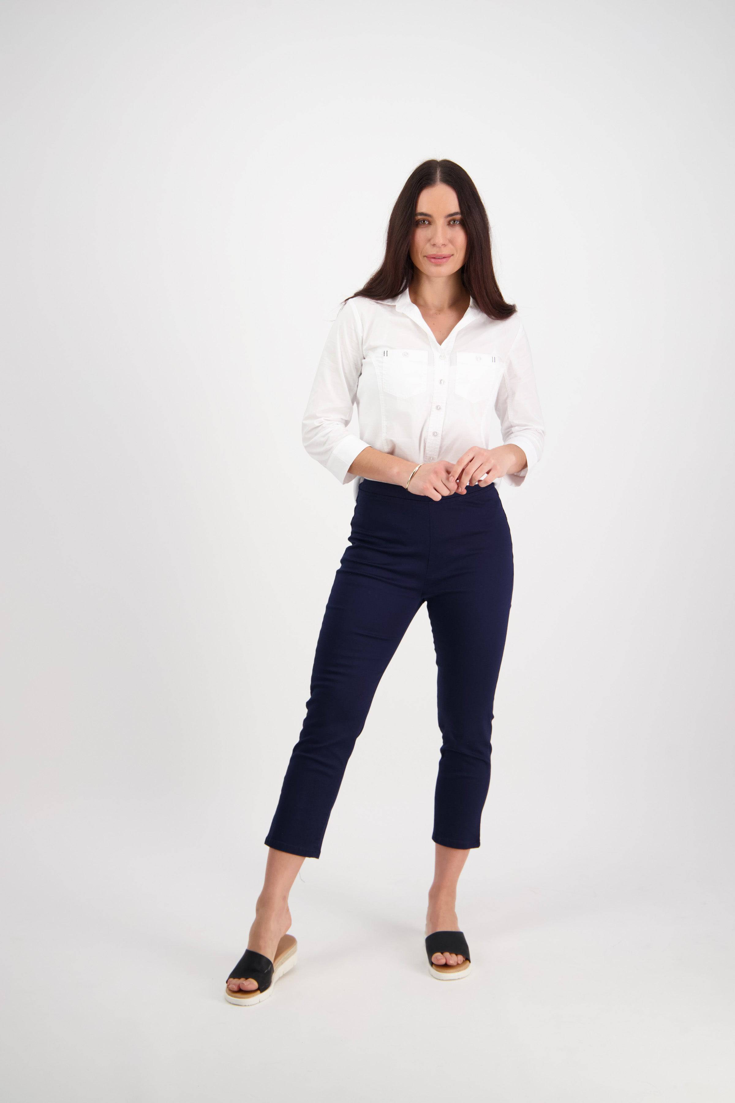 A woman with long dark hair poses confidently against a white background, wearing the Vassalli Harbour 7/8 Pant in Navy, a white button-up shirt, and black slip-on sandals.