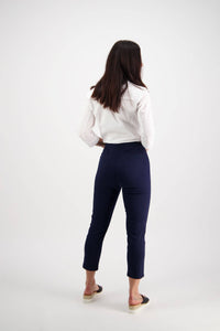 A woman stands facing away, wearing a white long-sleeve shirt and Vassalli Harbour 7/8 Pant in navy, featuring a high-waisted skinny fit, paired with black slip-on shoes with light soles, against a plain white background.