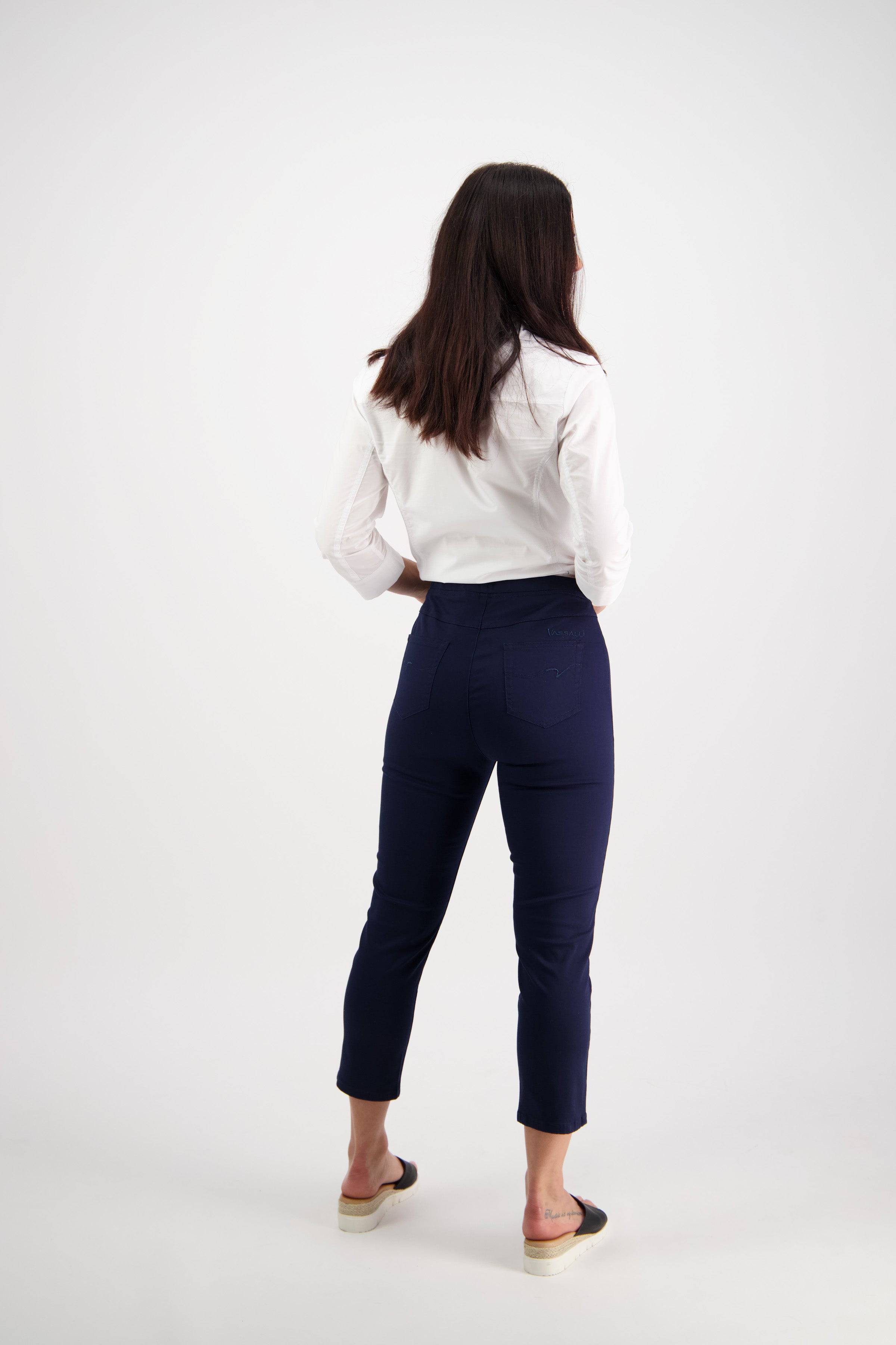 A woman stands facing away, wearing a white long-sleeve shirt and Vassalli Harbour 7/8 Pant in navy, featuring a high-waisted skinny fit, paired with black slip-on shoes with light soles, against a plain white background.