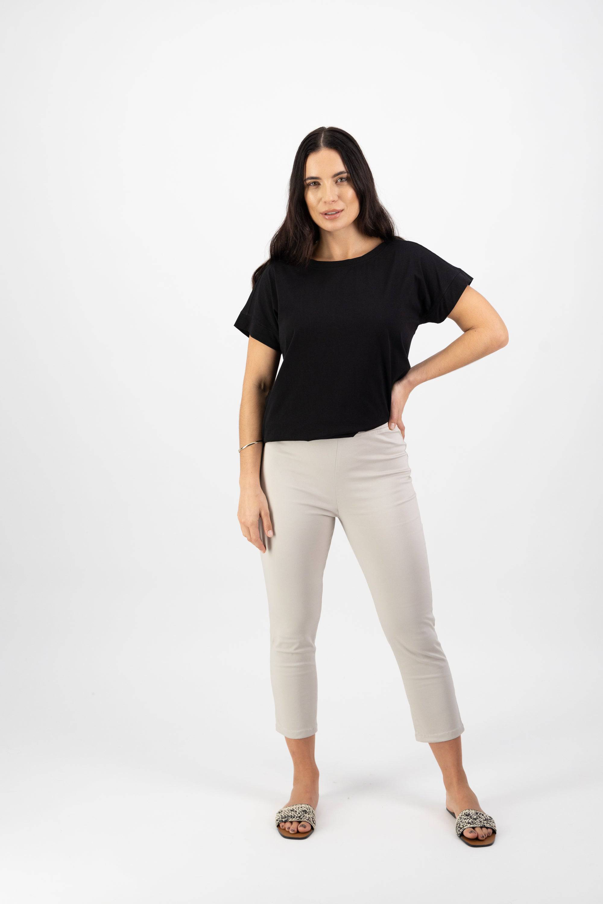 A woman with long dark hair stands against a white background, wearing a black short-sleeve top and Vassalli Harbour 7/8 Pant in Pumice. She completes the look with animal print slip-on shoes, one hand on her hip and a neutral expression.
