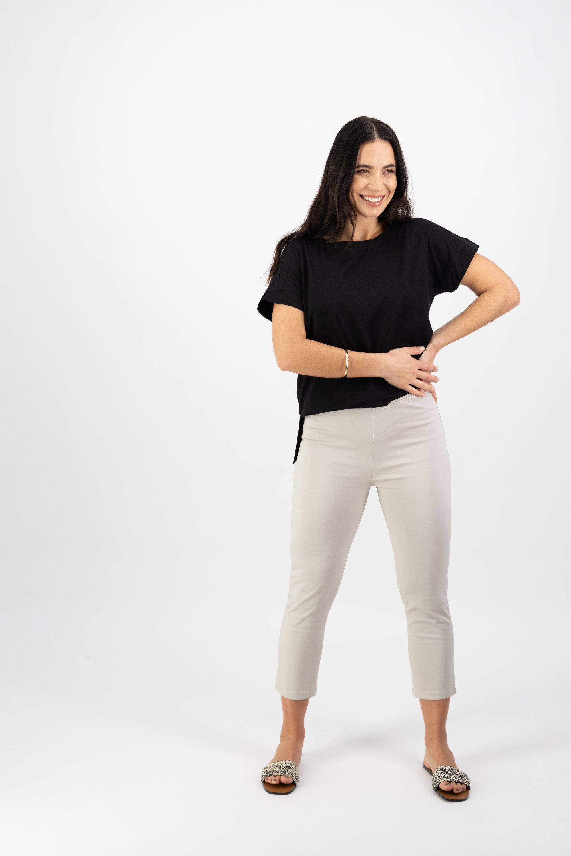 A woman with long dark hair smiles against a white background, wearing a black short-sleeve top and Vassalli Harbour 7/8 Pant in Pumice, paired with patterned sandals. One hand rests on her hip, the other across her waist.