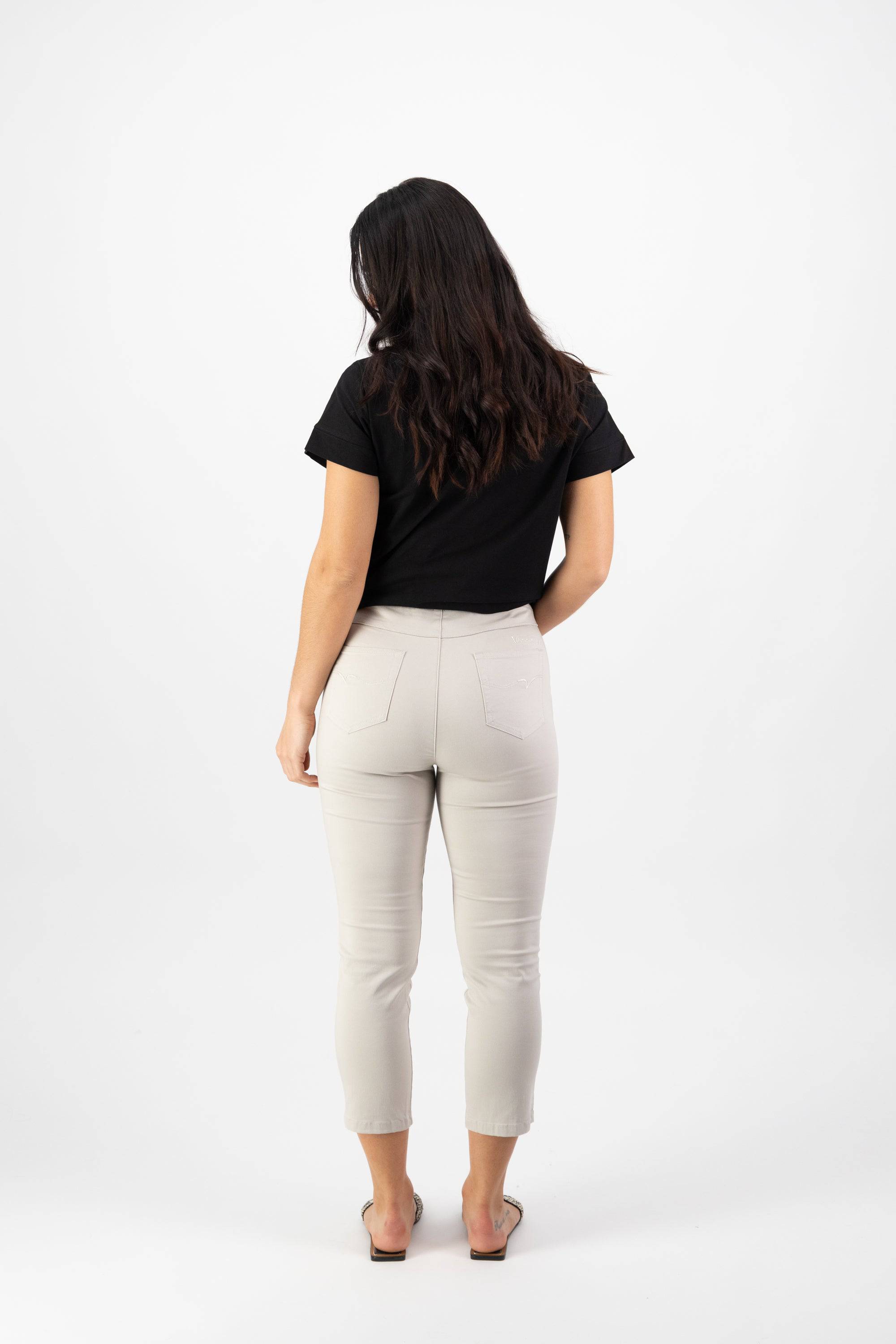 A woman with long dark hair faces away in a black short-sleeve top and Vassalli Harbour 7/8 Pant in Pumice, featuring pull-on stretch fabric skinny leg design, paired with flat shoes, set against a plain white background.