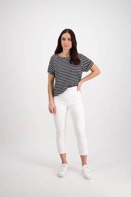 A woman with long dark hair wears the Vassalli Harbour 7/8 Pant in white, paired with a black and white striped top and white sneakers, posing against a plain white background with one hand on her hip.