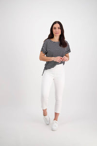 A woman with long dark hair and a neutral expression stands against a white background, wearing Vassalli's Harbour 7/8 Pant in white, paired with a black and white striped top and white sneakers, one foot slightly forward.
