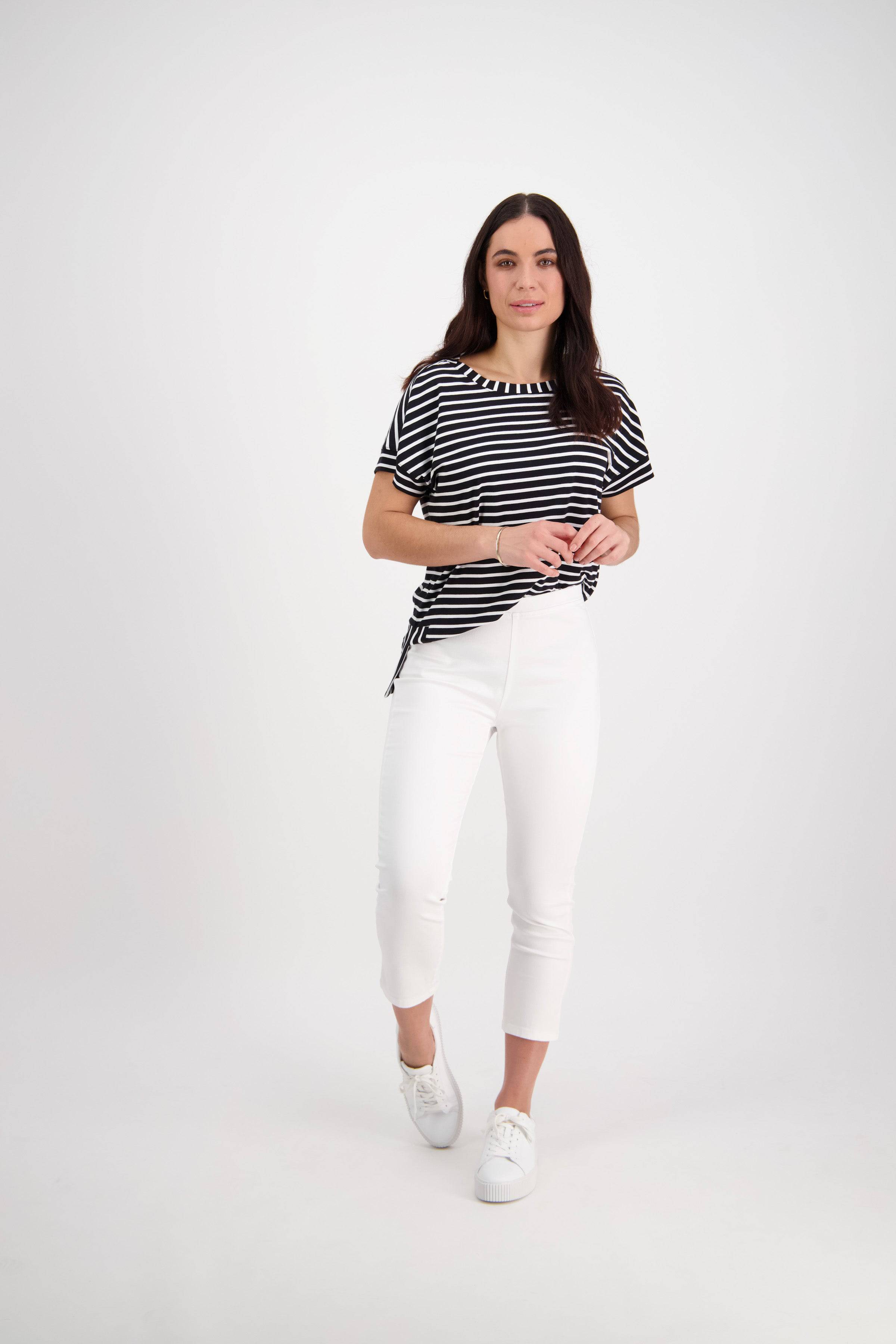 A woman with long dark hair and a neutral expression stands against a white background, wearing Vassalli's Harbour 7/8 Pant in white, paired with a black and white striped top and white sneakers, one foot slightly forward.