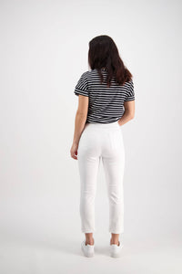 A woman with long dark hair stands facing away, wearing a black and white striped t-shirt, Vassalli Harbour 7/8 Pant in White, and white sneakers against a plain white background.