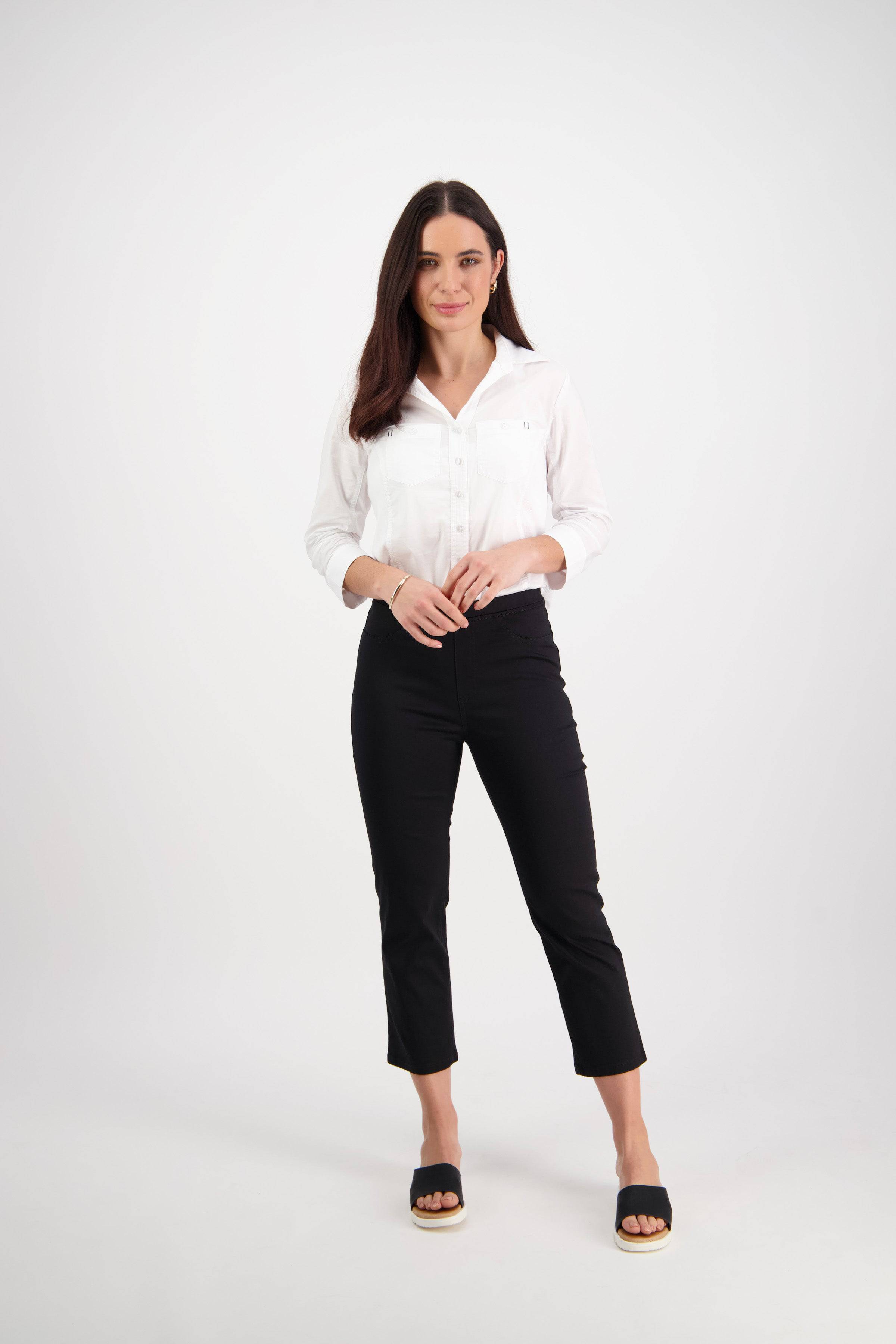 A woman with long dark hair stands facing forward, wearing a white button-up shirt and Vassalli Sierra 7/8 Pant in black with black sandals, against a plain white background.