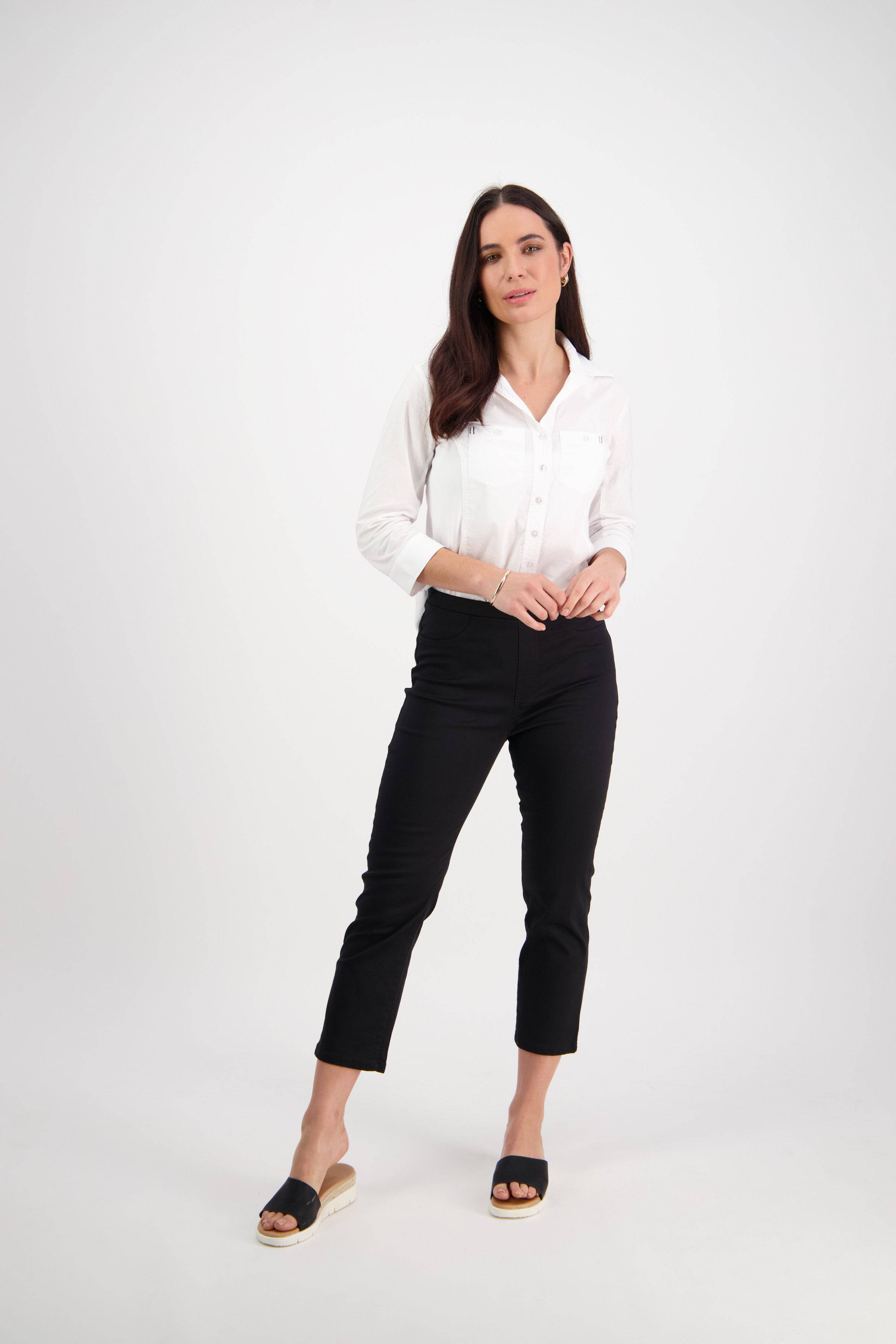 A woman with long dark hair poses confidently against a white background, wearing Vassalli's Sierra 7/8 Pant in Black with a white button-up shirt and black open-toe shoes, one hand in her pocket.