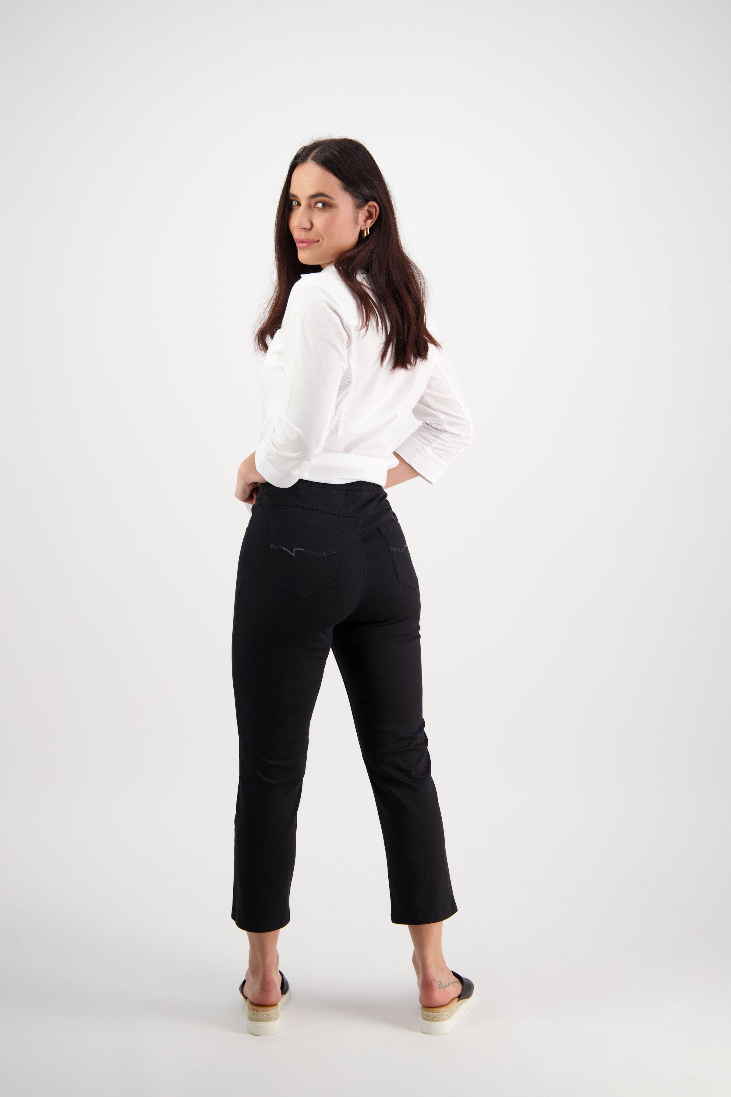 A woman with long dark hair stands in a white studio, wearing a white long-sleeve shirt, Vassalli Sierra 7/8 Pant in Black, and slip-on sandals as she turns slightly to look over her shoulder.