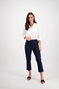 A woman with long brown hair poses in a studio, wearing the Vassalli Sierra 7/8 Pant in navy and a white button-up shirt with black slide sandals. She stands against a plain white background, one hand on her hip and a neutral expression.