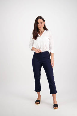 A woman with long brown hair poses in a studio, wearing the Vassalli Sierra 7/8 Pant in navy and a white button-up shirt with black slide sandals. She stands against a plain white background, one hand on her hip and a neutral expression.