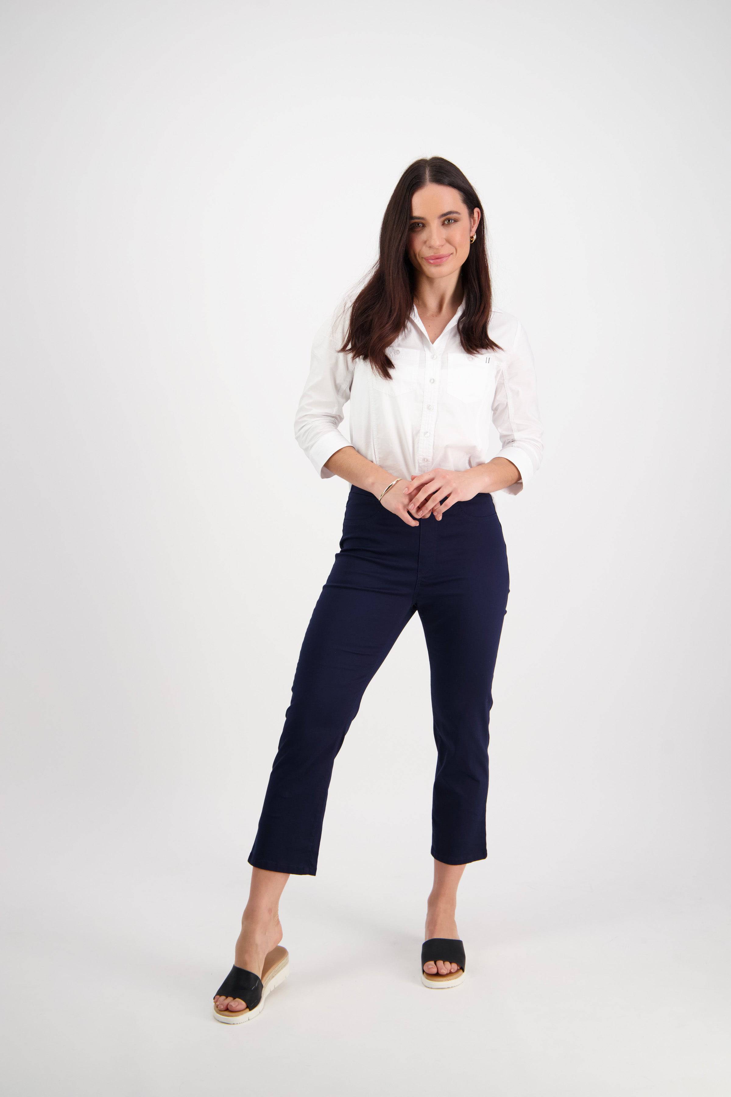 A woman with long dark hair stands against a white background, wearing a white shirt and Vassalli Sierra 7/8 Pant in Navy, paired with black open-toe sandals. She poses confidently and smiles slightly.