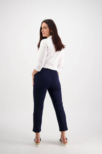 A woman with long dark hair looks over her shoulder in a white shirt and Vassalli Sierra 7/8 Pant - Navy, posing against a plain white background.