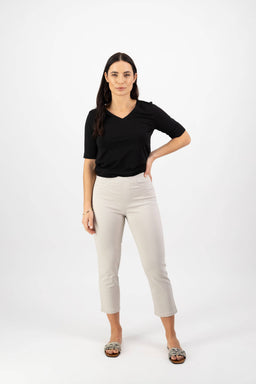 A woman with long dark hair stands against a white background, wearing a black short-sleeve top, patterned slip-ons, and Vassalli’s Sierra 7/8 Pant in Pumice. She faces forward with one hand on her hip.