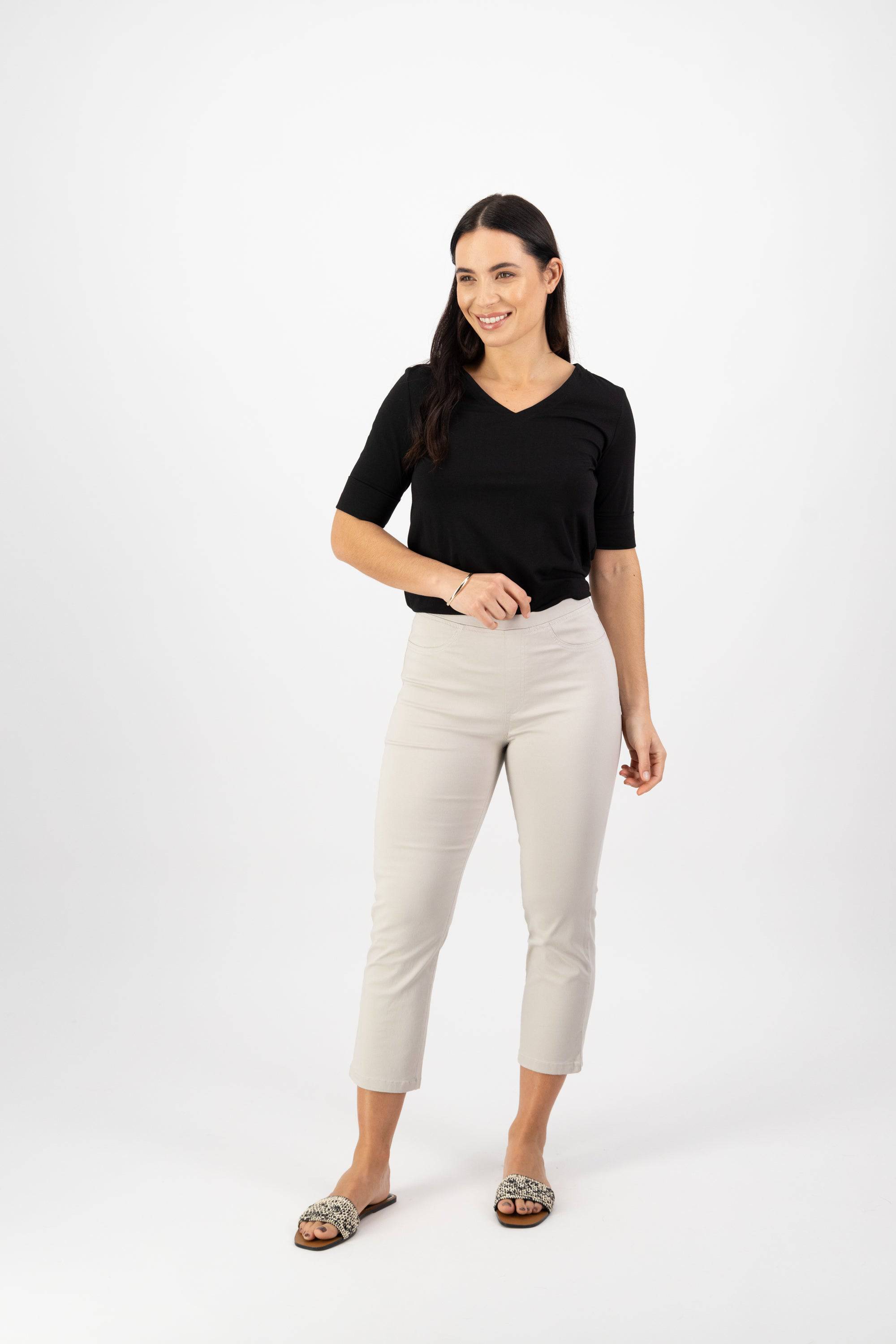 A woman with long dark hair wears a black V-neck top, Vassalli Sierra 7/8 Pant in Pumice, and slip-on shoes as she smiles against a white background, one hand relaxed by her side and the other slightly raised.