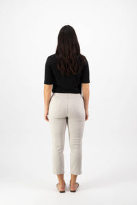 A woman with long, wavy dark hair faces away from the camera, wearing a black short-sleeve top, Vassalli Sierra 7/8 Pant in Pumice, and beige flat shoes against a plain white background.