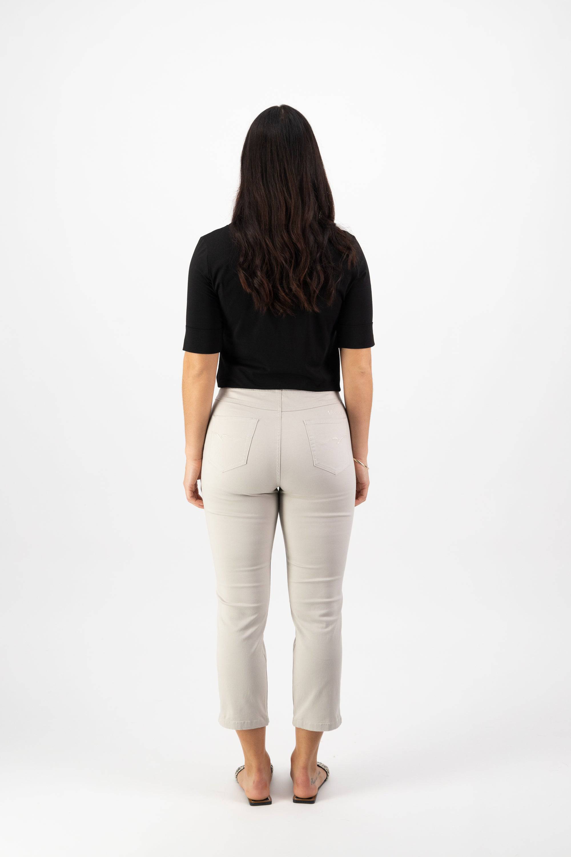 A woman with long, wavy dark hair faces away from the camera, wearing a black short-sleeve top, Vassalli Sierra 7/8 Pant in Pumice, and beige flat shoes against a plain white background.