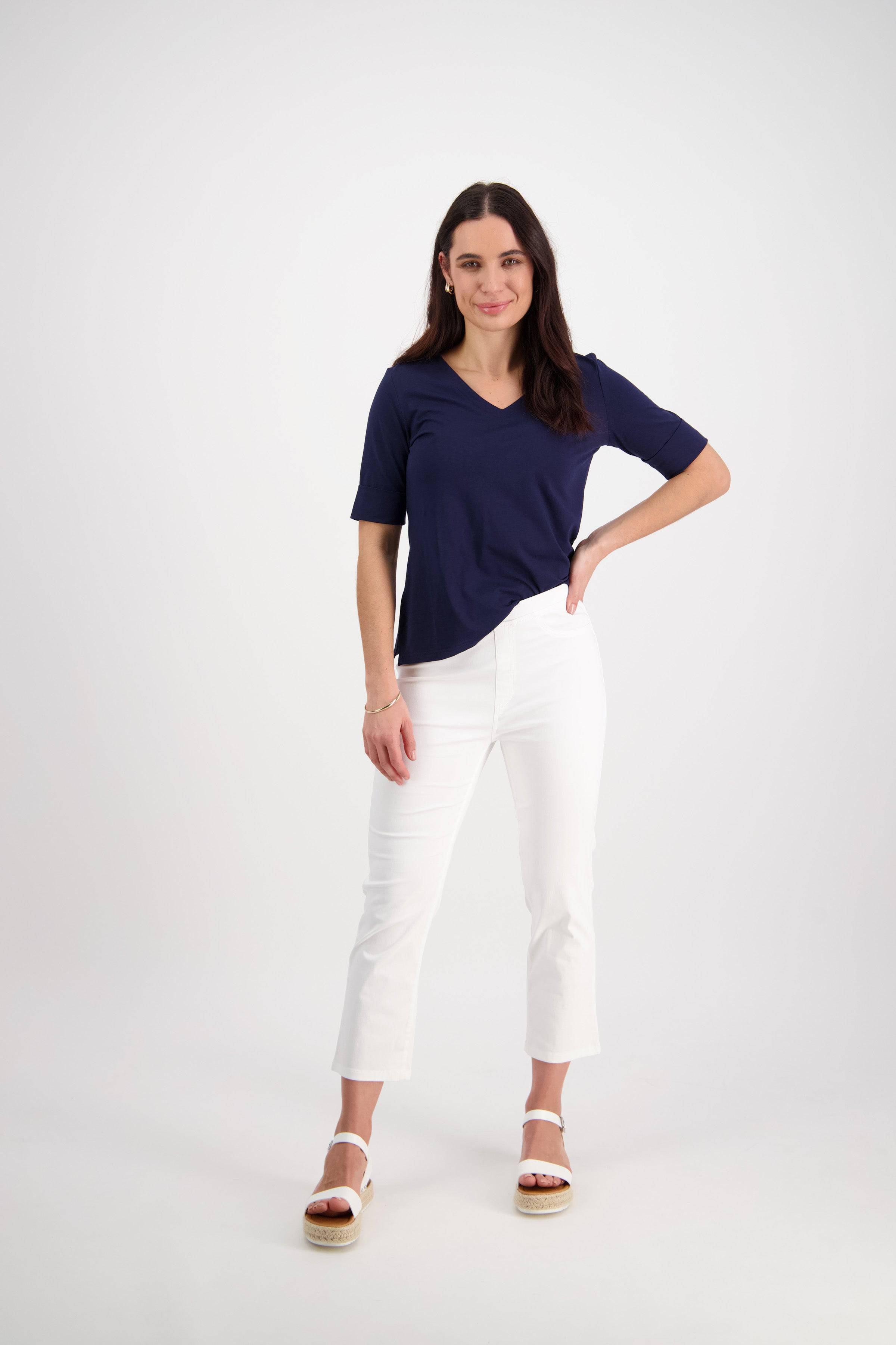 A woman stands against a plain white background, smiling. She wears a navy short-sleeve top, Vassalli Sierra 7/8 Pant in white, and white platform sandals. One hand rests on her hip while the other hangs at her side.