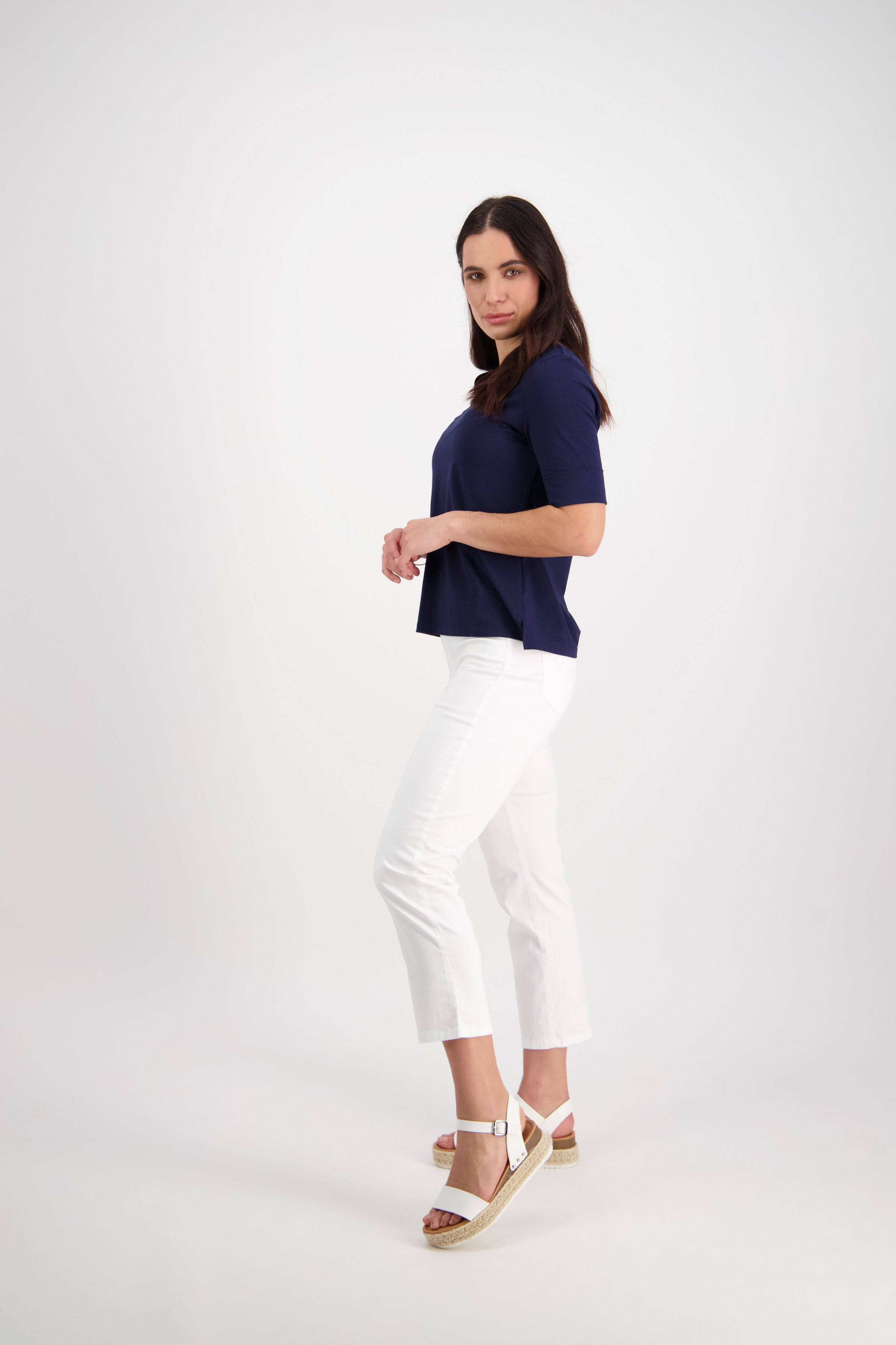 A woman stands in a studio against a white background, wearing the Vassalli Sierra 7/8 Pant in White with a navy top and white platform sandals, facing slightly sideways and looking at the camera with a neutral expression.
