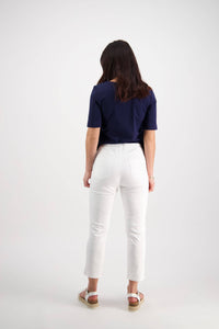 A woman with medium-length brown hair, facing away, wears a navy short-sleeve top and Vassalli Sierra 7/8 Pant in white, paired with white sandals, against a plain white background.