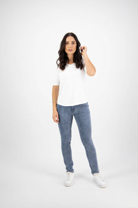 A woman with long dark hair stands against a white background, wearing blue jeans and the Vassalli Base Tee in white—a relaxed fit cotton jersey tee that completes her look. She faces the camera with one hand near her ear and a neutral expression.