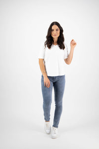 A woman with long, dark hair poses against a white background in a Vassalli Base Tee - White, paired with classic blue jeans and white sneakers. She looks at the camera, holding one hand up for a simple yet stylish look.