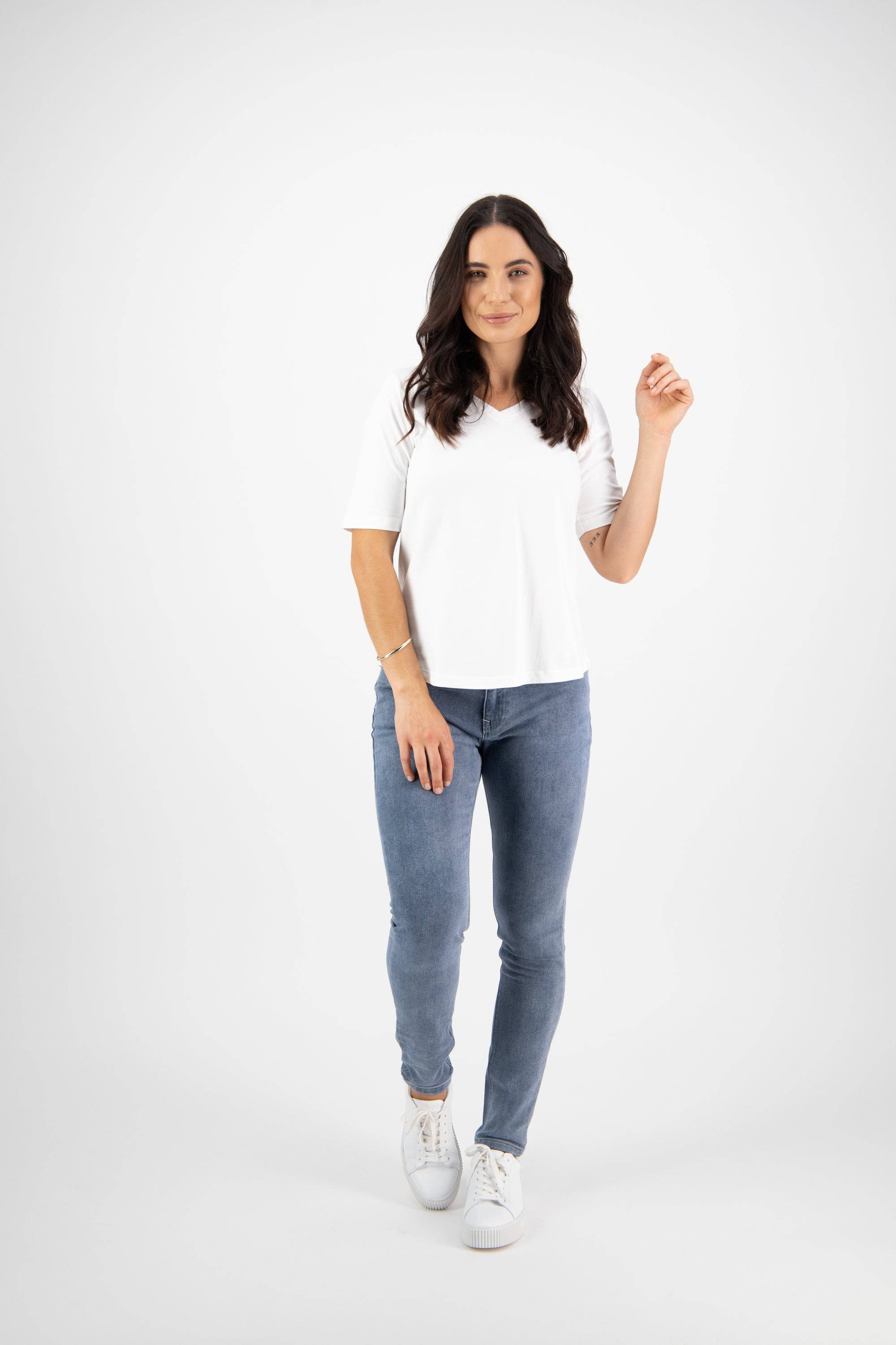 A woman with long, dark hair poses against a white background in a Vassalli Base Tee - White, paired with classic blue jeans and white sneakers. She looks at the camera, holding one hand up for a simple yet stylish look.