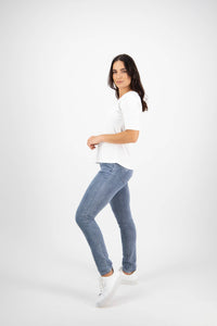 A woman with long dark hair stands sideways, looking at the camera. She wears the Vassalli Base Tee in white, styled with light blue jeans and white sneakers, posing against a plain white background.