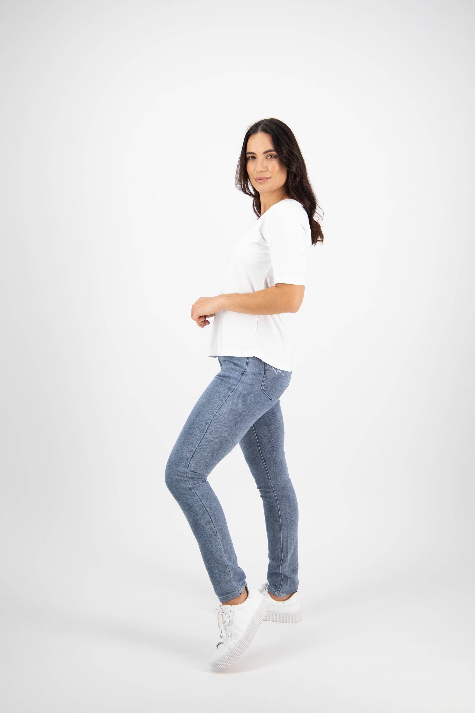 A woman with long dark hair stands sideways, looking at the camera. She wears the Vassalli Base Tee in white, styled with light blue jeans and white sneakers, posing against a plain white background.