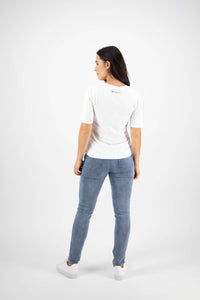 A woman stands with her back to the camera, wearing a Vassalli Base Tee in white, light blue jeans, and white sneakers against a plain white background.