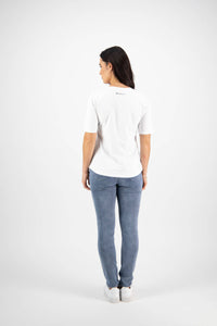 A woman with long dark hair faces away, wearing the Vassalli Base Tee in white and light blue jeans with white sneakers, set against a plain white background.